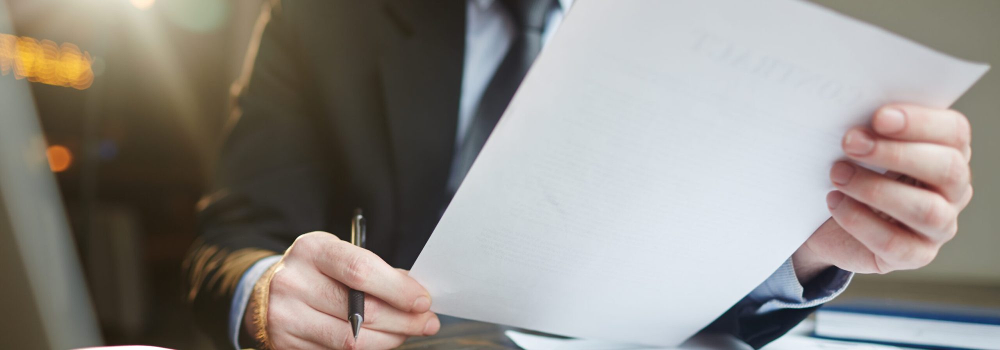 Portrait of modern bearded businessman holding papers in hands, reading and analyzing contract documentation at desk with laptop